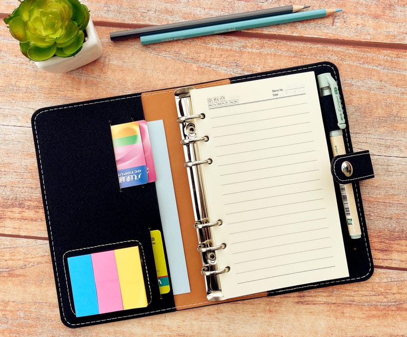 Op de afbeelding: Zwart leren planner met gelijnd papier, een pen en een pennenhouder. De planner heeft een drukknoopsluiting en een vak voor kaarten en plaknotities. Een kleine vetplant en potloden staan op de achtergrond.
