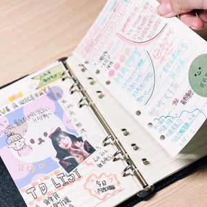 Op de afbeelding: Een open zwarte binder met gelijnde papieren pagina's, waarvan er &eacute;&eacute;n wordt opgetild om handgeschreven notities en kleurrijke stickers te onthullen. De linkerpagina bevat een "TD LIST" en een foto. De opgetilde pagina heeft de woorden "bottoms up" en "Reading Word."