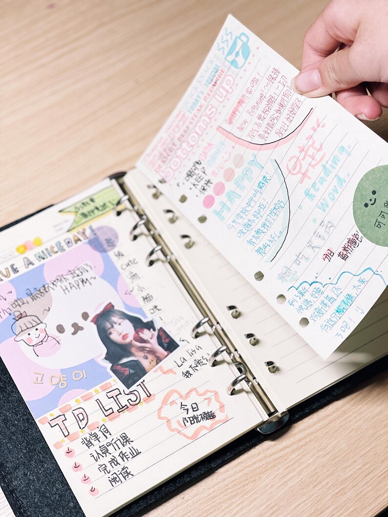 Op de afbeelding: Een open zwarte binder met gelijnde papieren pagina's, waarvan er &eacute;&eacute;n wordt opgetild om handgeschreven notities en kleurrijke stickers te onthullen. De linkerpagina bevat een "TD LIST" en een foto. De opgetilde pagina heeft de woorden "bottoms up" en "Reading Word."