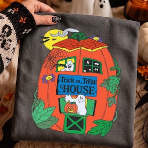 May include: A charcoal gray t-shirt featuring a graphic of a large orange pumpkin shaped like a house with a green roof. The design includes white ghosts, green vines, a yellow moon, and a blue sign with the text Trick or Treat HOUSE.