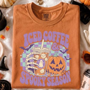 May include: A burnt orange crewneck t-shirt featuring a graphic of a skeleton hand holding an iced coffee cup next to a jack-o'-lantern. The text reads ICED COFFEE & SPOOKY SEASON in purple letters over a light purple and teal checkered background.