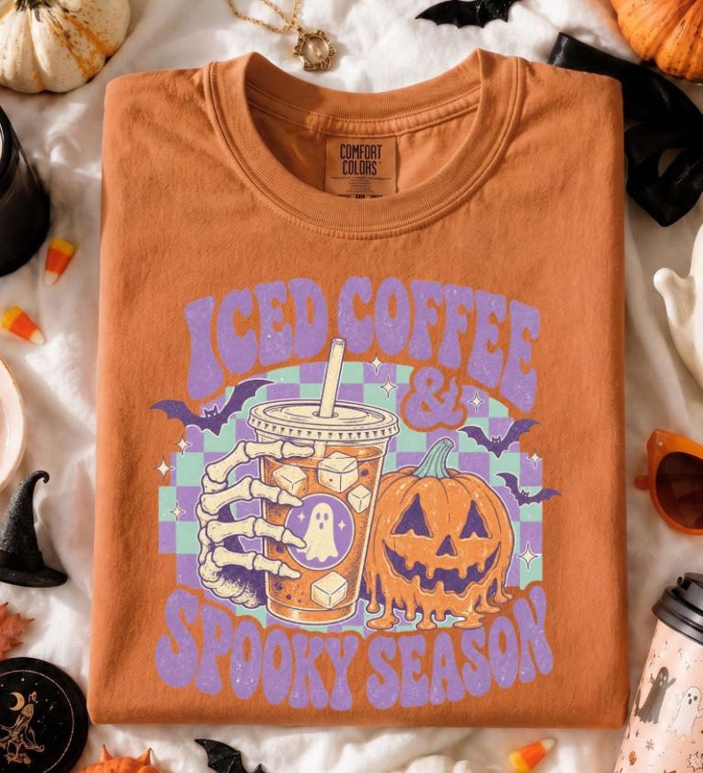 May include: A burnt orange crewneck t-shirt featuring a graphic of a skeleton hand holding an iced coffee cup next to a jack-o'-lantern. The text reads ICED COFFEE & SPOOKY SEASON in purple letters over a light purple and teal checkered background.