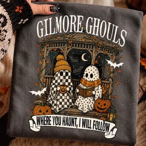 May include: A dark gray t-shirt featuring a graphic of two ghosts in a gazebo with autumn leaves, pumpkins, and bats. The text reads GILMORE GHOULS at the top and WHERE YOU HAUNT, I WILL FOLLOW on a banner at the bottom. The ghosts wear hats and scarves.