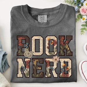 May include: A charcoal gray t-shirt featuring the text BOOK NERD in large block letters. The letters have a vintage book page background with colorful floral illustrations and a butterfly graphic integrated into the design.