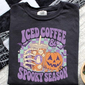 May include: A black t-shirt featuring a graphic of a skeleton hand holding an iced coffee cup with a ghost icon, next to a melting jack-o'-lantern. The design includes purple text reading "ICED COFFEE & SPOOKY SEASON" over a light green and purple checkered background.