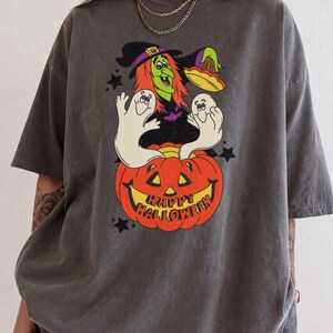 May include: A charcoal gray t-shirt featuring a graphic print of a green-skinned witch with orange hair, two white ghosts, and a large orange jack-o'-lantern. The pumpkin has a carved face and the text "HAPPY HALLOWEEN" printed in red.