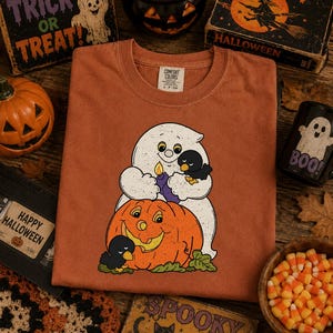 May include: A rust-colored t-shirt featuring a graphic of a white ghost holding a purple candle and a small black bird, sitting atop a large orange jack-o'-lantern with another black bird nearby. The shirt has a Comfort Colors brand neck label.