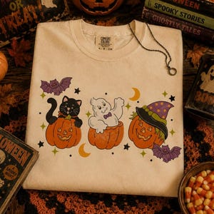 May include: A cream-colored t-shirt featuring a graphic print of a black cat in a pumpkin, a white ghost in a pumpkin, and a jack-o'-lantern wearing a witch hat. The design includes two purple bats, yellow crescent moons, and small black and green stars.