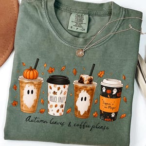 May include: A sage green t-shirt featuring a graphic of four coffee cups with autumn themes, including ghosts, pumpkins, and falling leaves. The text reads "HELLO, FALL!", "Sippin on Magic", and "Autumn leaves & coffee please". A silver necklace rests on the collar.