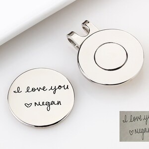 May include: Two silver-colored golf ball markers. One marker has the handwritten text "I love you Megan" with a heart symbol. The other marker has a clip for attaching to a hat or visor.