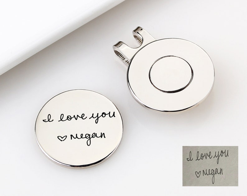 May include: Two silver-colored golf ball markers. One marker has the handwritten text "I love you Megan" with a heart symbol. The other marker has a clip for attaching to a hat or visor.