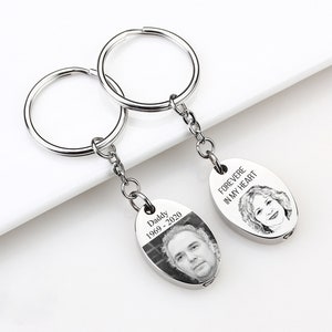 May include: Two silver keychains with oval pendants. One pendant has a photo and the text "Daddy 1969-2020". The other pendant has a portrait and the text "FOREVER IN MY HEART". The keychains are on a white surface.