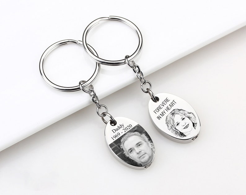 May include: Two silver keychains with oval pendants. One pendant has a photo and the text "Daddy 1969-2020". The other pendant has a portrait and the text "FOREVER IN MY HEART". The keychains are on a white surface.