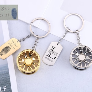 May include: Two keyrings with wheel-shaped charms, one gold-toned, the other silver. Each keyring has a tag with personalised text. One tag has a fingerprint and the text "My LV 02-05-23". The other tag says "LORENA 9.25.2023".