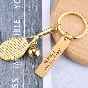 May include: A gold-colored tennis-themed keychain. The keychain features a tennis racket and ball charm, along with a rectangular tag engraved with "Jenny & David 7.19.2025". The keychain is attached to a round key ring.