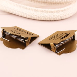 May include: Two bronze-colored graduation cap-shaped money clips with a spring mechanism. Each clip has text that reads "Class of 2023" and "Congratulations". The clips are on a light pink surface.