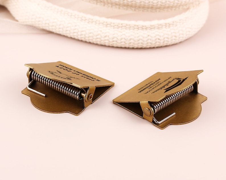 May include: Two bronze-colored graduation cap-shaped money clips with a spring mechanism. Each clip has text that reads "Class of 2023" and "Congratulations". The clips are on a light pink surface.