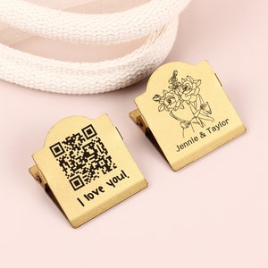 May include: Two gold-colored metal clips. One clip has a QR code and the text "I love you!". The other clip has a floral design and the names "Jennie & Taylor". Both clips have a hinged design.