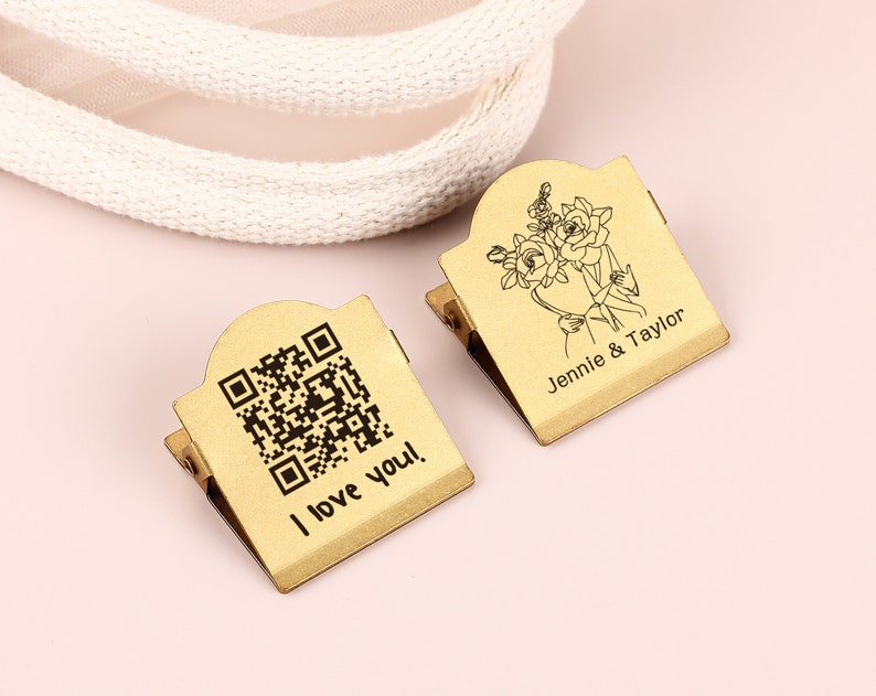 May include: Two gold-colored metal clips. One clip has a QR code and the text "I love you!". The other clip has a floral design and the names "Jennie & Taylor". Both clips have a hinged design.