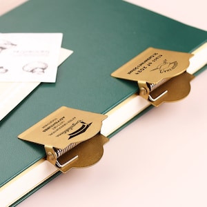 May include: Two brass-colored graduation cap-shaped book clips are attached to a green journal. The clips have a spring mechanism and text that reads "Congratulations, After Graduation, Let's Get Adventures Together." A card with mushroom illustrations is visible.