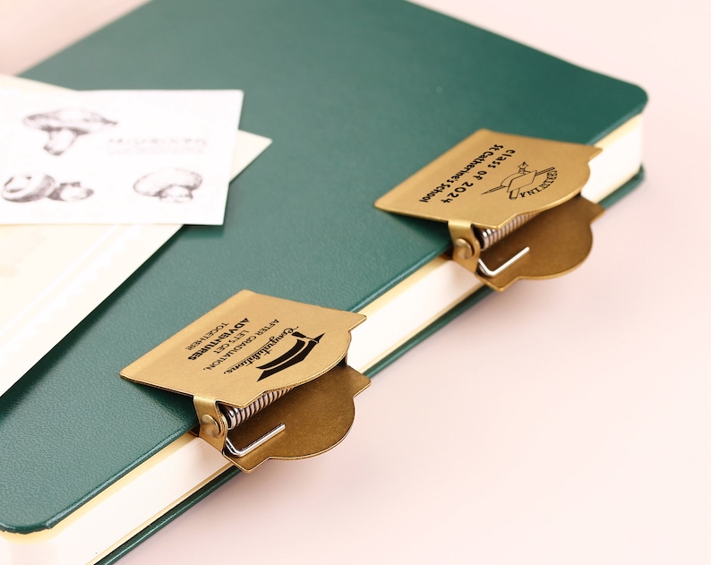 May include: Two brass-colored graduation cap-shaped book clips are attached to a green journal. The clips have a spring mechanism and text that reads "Congratulations, After Graduation, Let's Get Adventures Together." A card with mushroom illustrations is visible.
