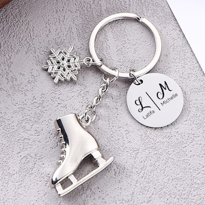 Op de afbeelding: Zilverkleurige sleutelhanger met een sneeuwvlokbedel, een figuurschaatsbedel en een ronde tag met de initialen "L | M" en de namen "Michelle" en "Latifa".