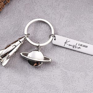 Op de afbeelding: Een zilverkleurige sleutelhanger met een raket, een planeet met ringen en een witte rechthoekige tag. De tag is gegraveerd met de naam "Kenisha" en de datum "3.25.2025". De sleutelhanger ligt op een grijze ondergrond.