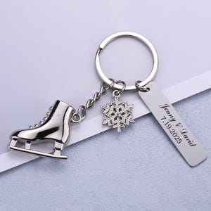 Op de afbeelding: Zilverkleurige sleutelhanger met een miniatuur schaats, een sneeuwvlok bedel en een rechthoekig label. Het label is gegraveerd met de namen "Jenny & David" en de datum "7.19.2025". De sleutelhanger ligt op een witte ondergrond.
