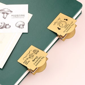 May include: A green notebook with a white-edged cover, featuring two gold-colored metal clips. One clip reads "The moment you became my Daddy" with a clock and footprints. The other clip reads "The moment I became a Daddy" with baby details. A card with mushroom illustrations is also present.