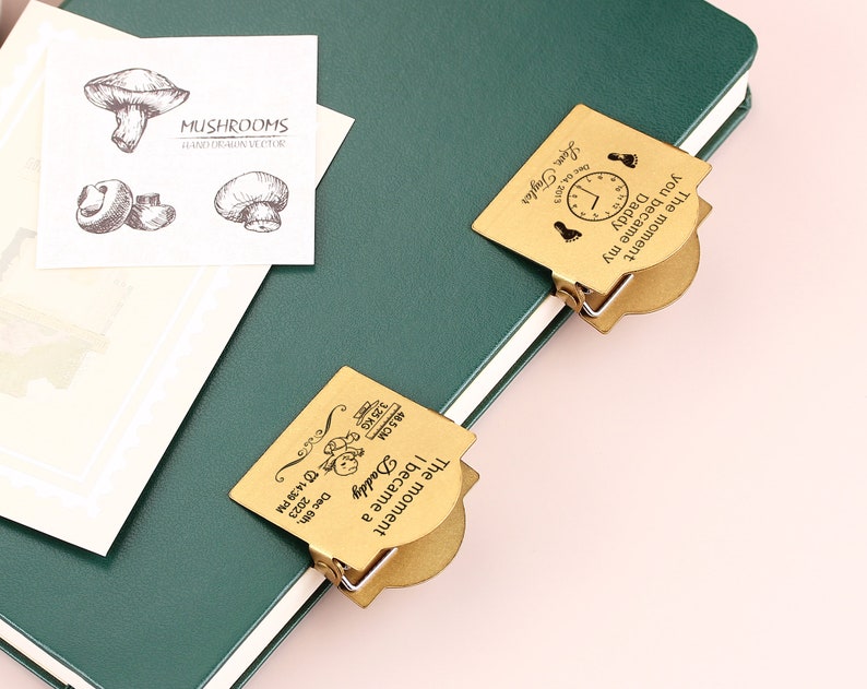 May include: A green notebook with a white-edged cover, featuring two gold-colored metal clips. One clip reads "The moment you became my Daddy" with a clock and footprints. The other clip reads "The moment I became a Daddy" with baby details. A card with mushroom illustrations is also present.