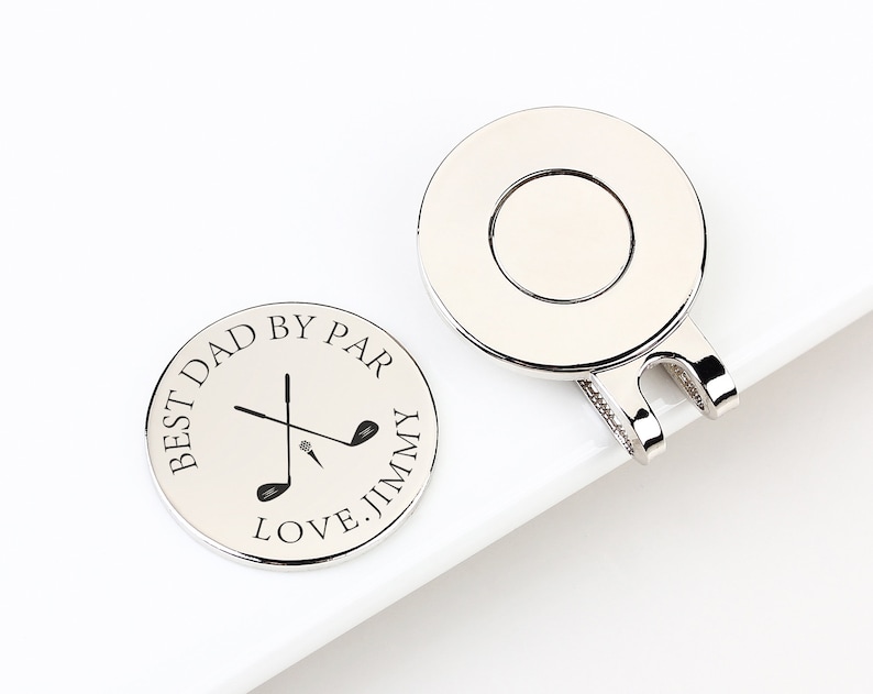 May include: Two silver golf ball markers. One marker has the text "BEST DAD BY PAR LOVE. JIMMY" around a golf club graphic. The other marker has a circular design with a clip. Golf-themed gift.