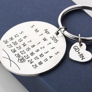 Op de afbeelding: Zilveren sleutelhanger met een ronde hanger met een kalender met de datum 30 april 2015 en een hartvormige hanger met de tekst "GD+RN".