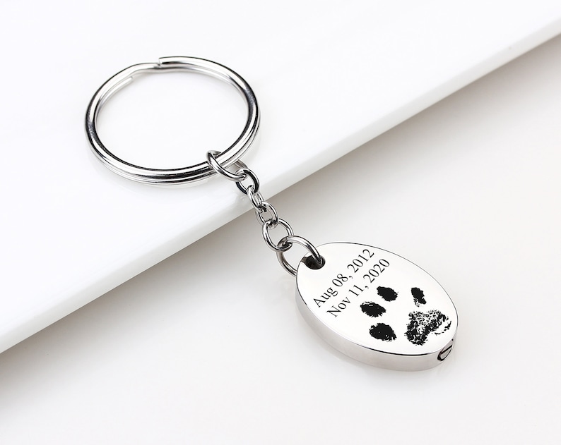 May include: A silver-colored keychain with a paw print design and the dates Aug 08, 2012 and Nov 11, 2020 engraved on an oval pendant. The keychain has a circular ring and a chain.