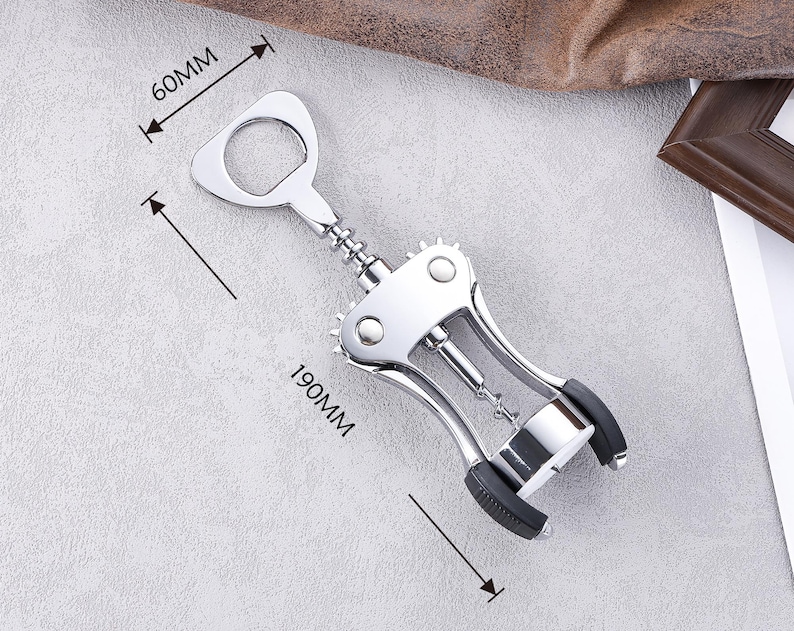 K&ouml;nnte beinhalten: Ein polierter Wein&ouml;ffner aus Chrom mit einem Flaschen&ouml;ffner, der 19 cm lang und 6 cm breit ist. Der &Ouml;ffner hat ein Hebeldesign mit schwarzen Gummi-Griffen und einem Korkenzieher. Das Werkzeug dient zum &Ouml;ffnen von Weinflaschen.