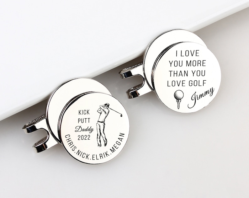 May include: Two silver golf ball markers with a magnetic clip. One marker has the text "I LOVE YOU MORE THAN YOU LOVE GOLF" and "Jimmy". The other marker has "KICK PUTT Daddy 2022" and names.