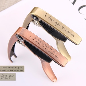 May include: Two guitar capo clamps with engraved messages. The top capo is gold with the message "i love you 2-23-20". The bottom capo is copper with the message "I believe in you. I'm here if you need me."