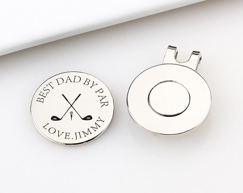 May include: Two silver golf ball markers. One marker has the text "BEST DAD BY PAR LOVE. JIMMY" around a crossed golf club and tee design. The other marker has a magnetic clip on the back. Golf-themed gift.