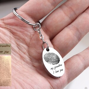 May include: Silver-colored keychain with an oval pendant featuring a fingerprint and the inscription "my grandma I love you." The keychain includes a key ring and a small chain. A separate card displays the fingerprint and text.