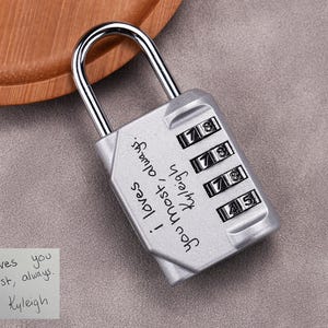 May include: A silver combination lock with a shiny metal shackle. The lock face has four number dials and the words "i loves you most, always. Kyleigh" are written on the side. A small note with the same text is next to the lock.