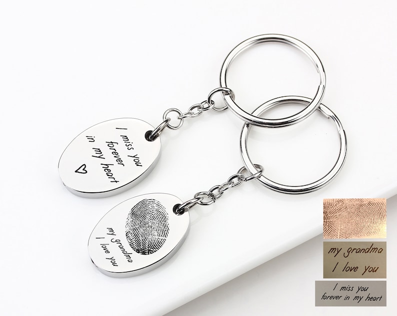 May include: Two silver keychains with oval pendants. One pendant has a fingerprint and the text "my grandma I love you." The other pendant has the text "I miss you forever in my heart" and a heart symbol. The keychains are connected by a chain and a keyring.