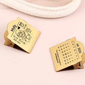 May include: Two gold-colored metal clips. One clip features a calendar for July 2023. The other clip has a baby-themed design with the name "Molly", birth details, and a baby illustration. The clips are on a pink surface.