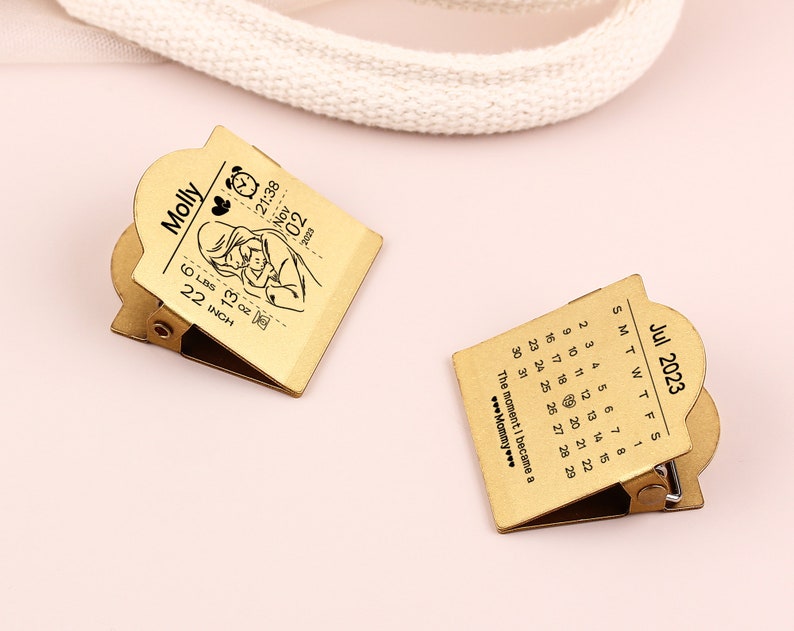 May include: Two gold-colored metal clips. One clip features a calendar for July 2023. The other clip has a baby-themed design with the name "Molly", birth details, and a baby illustration. The clips are on a pink surface.
