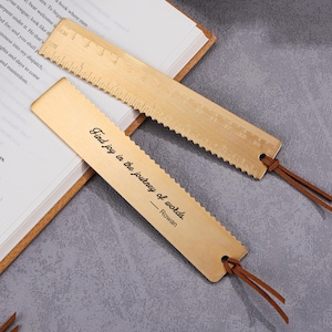 Può includere: Due segnalibri in ottone con segni di righello e un bordo decorativo sono appoggiati su un libro aperto. Un segnalibro ha la citazione "Find joy in the journey of words - Rowan" e lacci in pelle.
