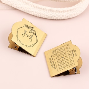 May include: Two gold-colored metal clips with a calendar and personalized text. One clip displays a July 2023 calendar with a heart on the 19th and the text "The day you become my boyfriend!". The other clip has the names "Smith & Julia" and a floral design.