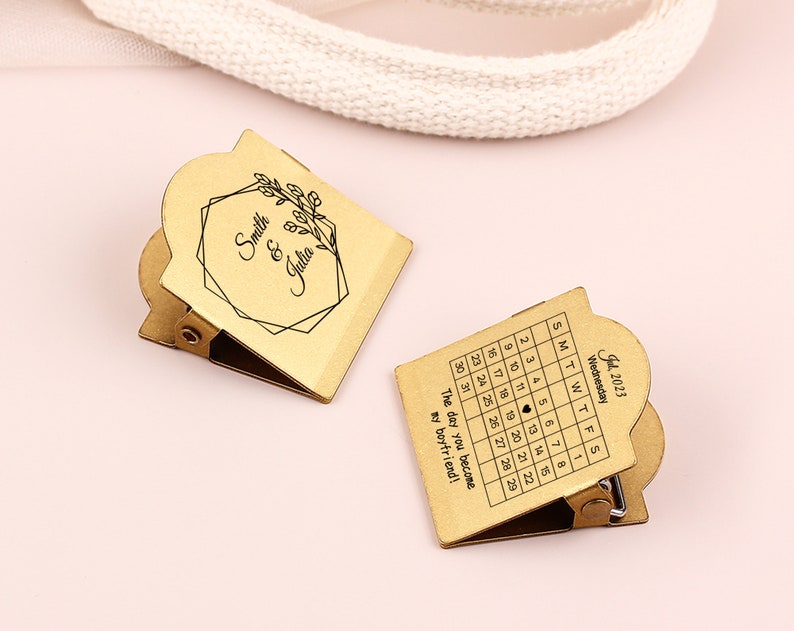 May include: Two gold-colored metal clips with a calendar and personalized text. One clip displays a July 2023 calendar with a heart on the 19th and the text "The day you become my boyfriend!". The other clip has the names "Smith & Julia" and a floral design.