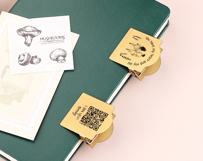 May include: A green notebook with two brass-colored page clips. One clip has the text "You have my heart and my ass" and a drawing. The other clip has a QR code and the text "I love you! @mags". A card with mushroom illustrations is also present.