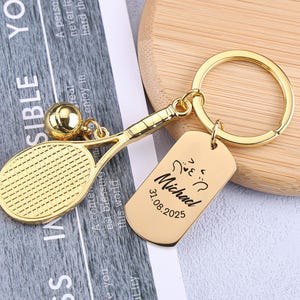 Peut inclure: Un porte-clés doré en forme de raquette de tennis avec un charm en forme de balle et une étiquette rectangulaire. L'étiquette est gravée du nom "Michael" et de la date "31.08.2025".