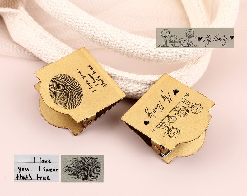 May include: Two gold-colored metal clips. One clip features a fingerprint and the text "I love you. I swear that's true." The other clip has a family illustration and the words "My Family." A white rope is in the background.