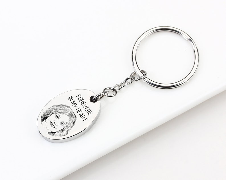 May include: A silver-colored keychain with an oval pendant. The pendant features a portrait and the text "FOREVERE IN MY HEART". The keychain includes a chain and a large ring for keys.