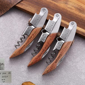 May include: Three personalized wine bottle openers with wooden handles and stainless steel accents. Each opener has a corkscrew and a small blade. One opener is engraved with "I love you so much Suzy", another with "Georgia", and the third with a date.